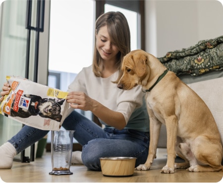 Egy nő Hill's Pet Nutrition állateledelt önt egy mérőedénybe, miközben egy kutya figyelmesen nézi